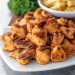 Close-up of grilled chicken nuggets with crispy char marks served on a white plate beside fries